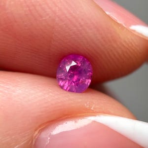 Pode incluir: Uma pedra preciosa rosa choque, facetada e de formato oval. A gema tem um corte brilhante com múltiplas facetas que refletem a luz. A pedra é segurada entre dois dedos, com um fundo neutro.