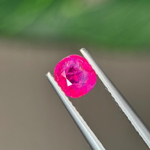 Puede incluir: Una gema de color rosa intenso con corte tipo cojín, sostenida por unas pinzas de metal plateado. La piedra presenta una superficie facetada que refleja la luz, mostrando un tono magenta saturado con inclusiones internas y bordes definidos.