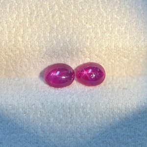 May include: Two oval-shaped, cabochon-cut star rubies with a deep magenta-pink hue. Each gemstone displays a distinct six-rayed star effect, known as asterism, visible on the smooth, polished, and domed surface of these two loose gemstones.