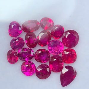 Puede incluir: Una colección de gemas de rubí rojo intenso facetadas en varias formas, incluyendo ovaladas, redondas y en forma de pera. Las gemas están dispuestas juntas, mostrando su corte brillante y su rico color.