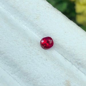 Może przedstawiać: Fasetowany, owalny, głęboko czerwony kamień szlachetny. Kamień jest osadzony na białym, teksturowanym tle, z rozmytym zielono-żółtym tłem. Fasetki kamienia odbijają światło.