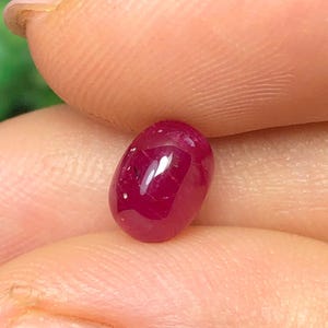Puede incluir: Una pequeña piedra preciosa de color rojo intenso y forma ovalada con una superficie cabujón lisa y pulida. La piedra muestra una calidad translúcida con sutiles inclusiones internas y un reflejo brillante en su parte superior redondeada, exhibiendo un tono carmesí rico.