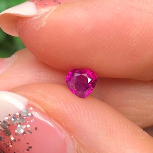 May include: A heart-shaped, faceted, deep pink gemstone. The gem is held between fingers, showcasing its brilliant cut and vibrant colour. The gemstone's facets catch the light, creating a sparkling effect. The background is blurred, emphasising the gem's details.