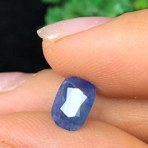 May include: A faceted, rectangular blue gemstone with a bright, reflective surface. The gem is held between fingers, showcasing its color and cut. The background is blurred, with hints of green foliage.