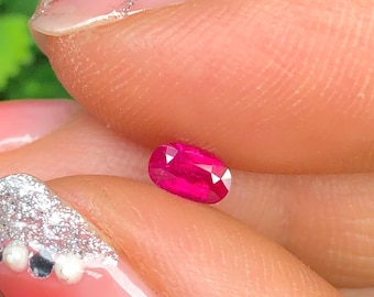 May include: A vibrant, oval-cut ruby gemstone. The ruby is a deep, saturated red color, with visible facets that catch the light. The gemstone is held between fingers, showcasing its size and brilliance. The background is blurred, highlighting the ruby's color and clarity.