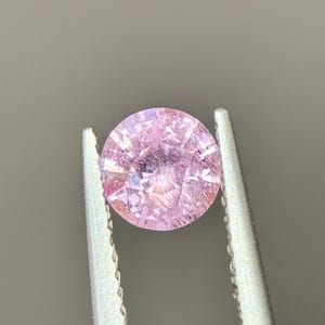May include: A round, pink gemstone with a faceted cut. The gemstone is held in tweezers.
