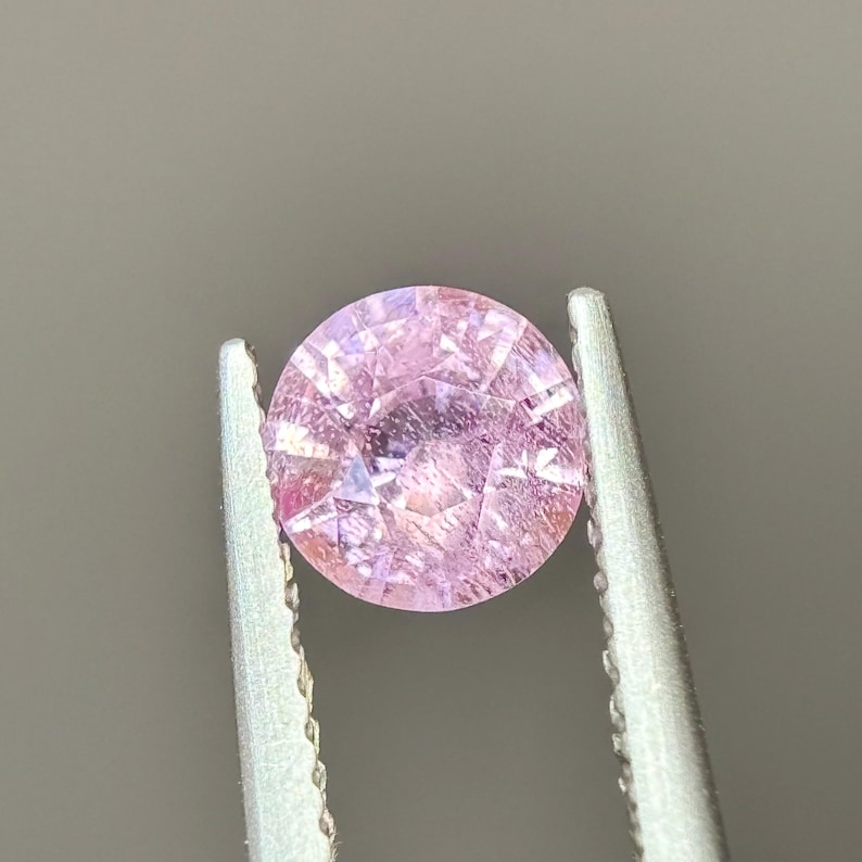 May include: A round, pink gemstone with a faceted cut. The gemstone is held in tweezers.