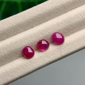 May include: Three polished, oval-shaped, deep red gemstones. The gems are displayed on a light beige, textured surface. The stones have a high lustre, reflecting light, and are likely rubies.