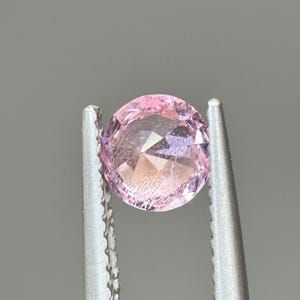 May include: A pink oval-shaped gemstone with a faceted cut, held in tweezers.