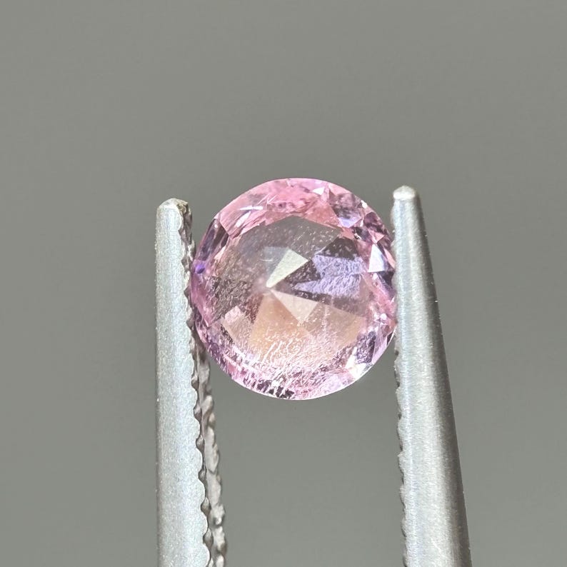 May include: A pink oval-shaped gemstone with a faceted cut, held in tweezers.