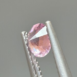May include: A pink gemstone with a faceted cut, held in tweezers. The gemstone is clear and has a slight pink hue.