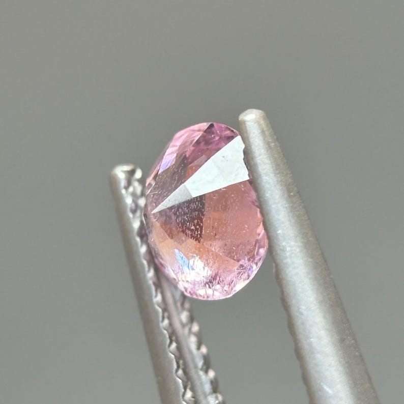 May include: A pink gemstone with a faceted cut, held in tweezers. The gemstone is clear and has a slight pink hue.