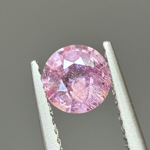 May include: A pink oval-shaped gemstone with a faceted cut. The gemstone is held in a pair of tweezers.