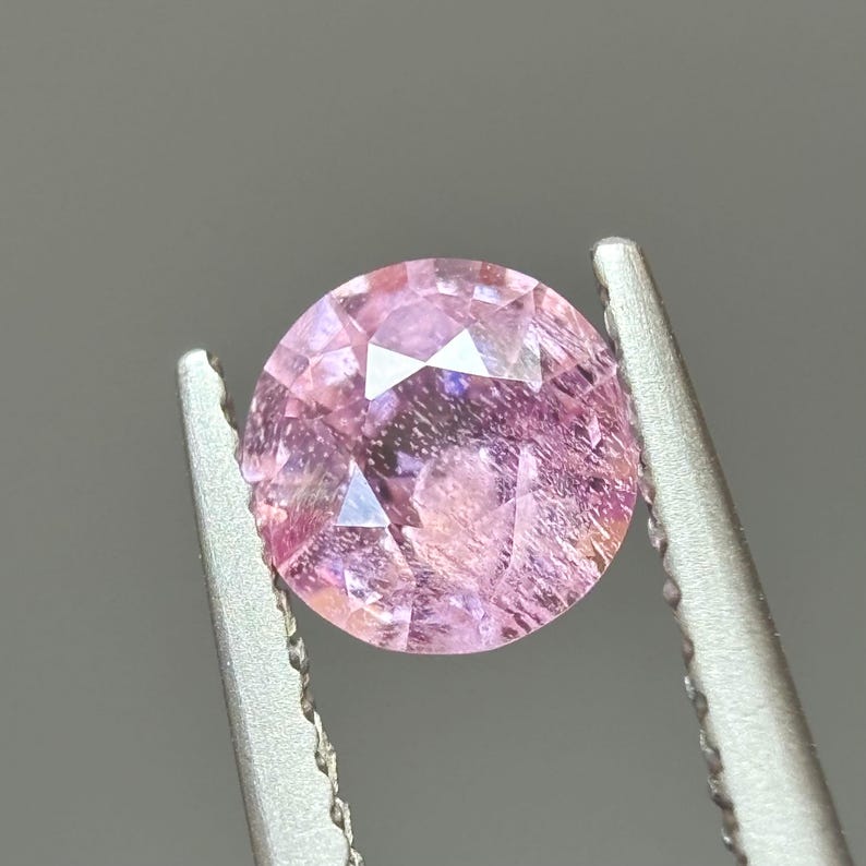 May include: A pink oval-shaped gemstone with a faceted cut. The gemstone is held in a pair of tweezers.