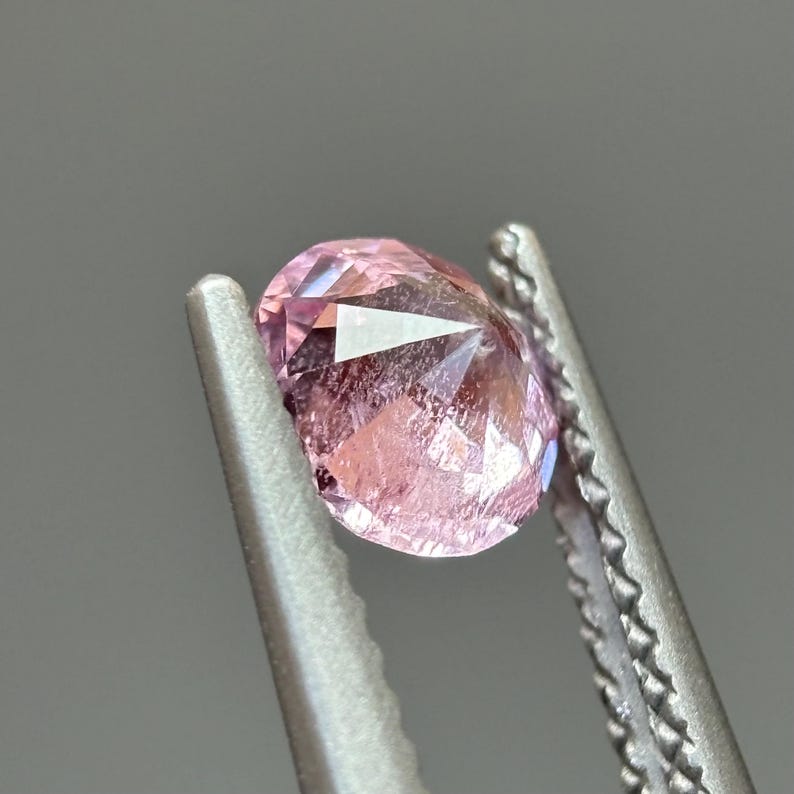 May include: A pink gemstone with a faceted cut, held in tweezers. The gemstone is oval shaped and has a translucent appearance.