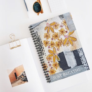 May include: A spiral-bound notebook with a floral design. The cover features yellow flowers with pink centers on a textured background with the words "JUST BREATHE". Other items include sunglasses, a paper clip, and a notepad.