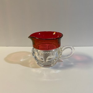 Puede incluir: Una jarra de cristal transparente con borde y asa de color rojo rubí. La jarra tiene un patrón decorativo de cristal tallado en la parte inferior. El asa es transparente y redondeada. La jarra está diseñada para servir líquidos.