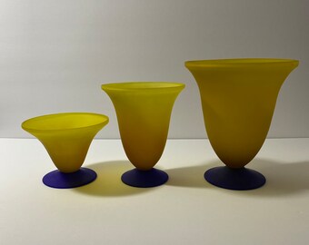 May include: Three yellow glass vases with a flared top and a dark blue base. The vases are graduated in size, with the smallest on the left and the largest on the right. The vases have a smooth, matte finish.