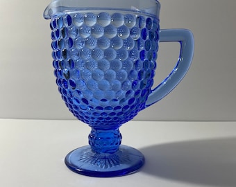 May include: A blue glass pitcher with a textured, bubble-like pattern. The pitcher has a handle and a spout for pouring. It sits on a round base with a decorative design. The pitcher is a vibrant shade of blue.