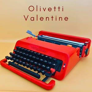 Peut inclure: Une machine à écrire Olivetti Valentine rouge vif avec des touches noires et un chariot argenté. Le texte "Olivetti Valentine" est affiché en rouge en haut. La machine à écrire est posée sur un fond orange clair.