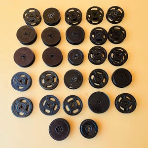 May include: A collection of twenty-seven black plastic film reels arranged in a grid. The reels vary in size and design, with some featuring open-spoke patterns and others having solid circular centers. Each reel has a central hole for mounting.