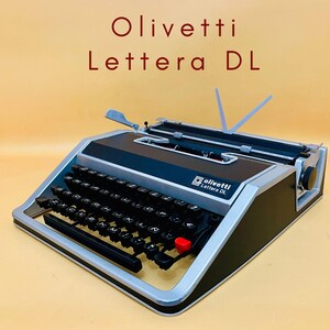 May include: Olivetti Lettera DL typewriter with black keys, a red key, and silver and black casing. The typewriter has a sleek design with a retro aesthetic. The text "Olivetti Lettera DL" is displayed in red at the top.