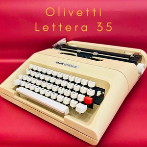 Może przedstawiać: Kremowa maszyna do pisania Olivetti Lettera 35 z białymi klawiszami i czerwonym klawiszem. Tekst "Olivetti Lettera 35" jest złotymi literami nad maszyną. Na czerwonym tle.