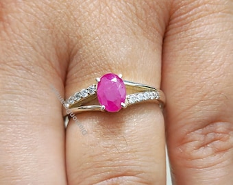 May include: A silver ring featuring a pink oval gemstone surrounded by smaller clear gemstones. The ring is worn on a finger.
