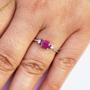 Peut inclure: Une bague en argent avec une pierre ovale rouge et deux petites pierres blanches. La bague est portée sur un doigt.