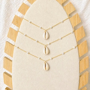 May include: Three gold-colored necklaces with a cowrie shell pendant. Each necklace has small white beads and a delicate chain. The necklaces are displayed on a cream-colored surface with a wooden decorative stand.