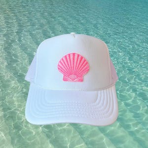 May include: White trucker hat with a pink seashell patch. The hat features a white front panel and bill, with a pink seashell design. The back of the hat is white mesh. A stylish accessory for beach or summer wear.