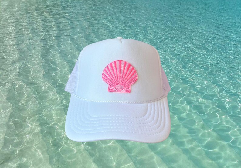 May include: White trucker hat with a pink seashell patch. The hat features a white front panel and bill, with a pink seashell design. The back of the hat is white mesh. A stylish accessory for beach or summer wear.