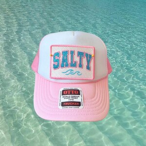 May include: A pink and white trucker hat with the word "SALTY" in a turquoise, glittery font on a rectangular patch. The hat has a pink brim and a white mesh back. The hat also has a label that says "OTTO" and "TRUCKER".
