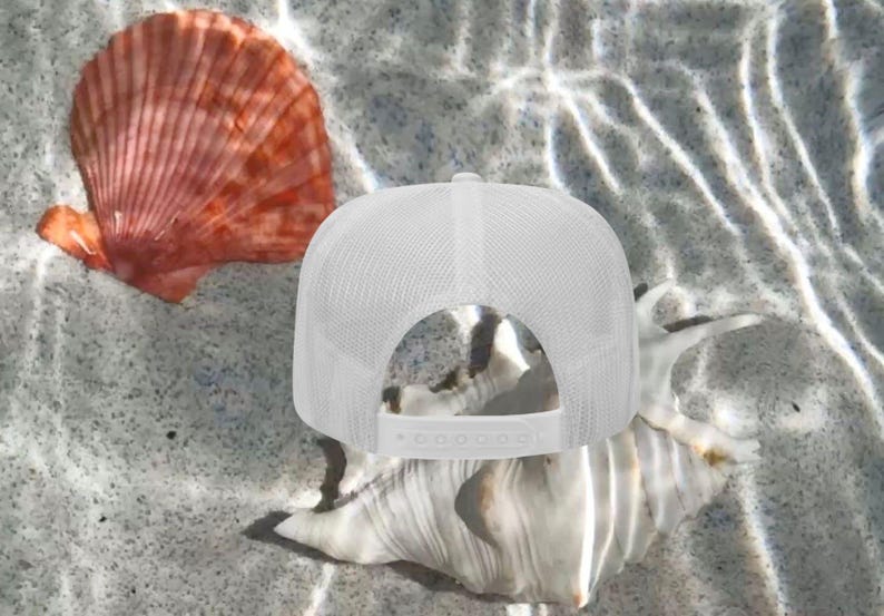 May include: A white trucker hat with a mesh back and adjustable snap closure, resting on a seashell. A vibrant orange seashell is also visible. The image is set against a sandy, watery background.