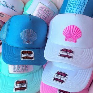 May include: A collection of trucker hats in various colors, including blue, white, and pink. Each hat features a seashell design on the front. The hats have a mesh back and a snap closure.