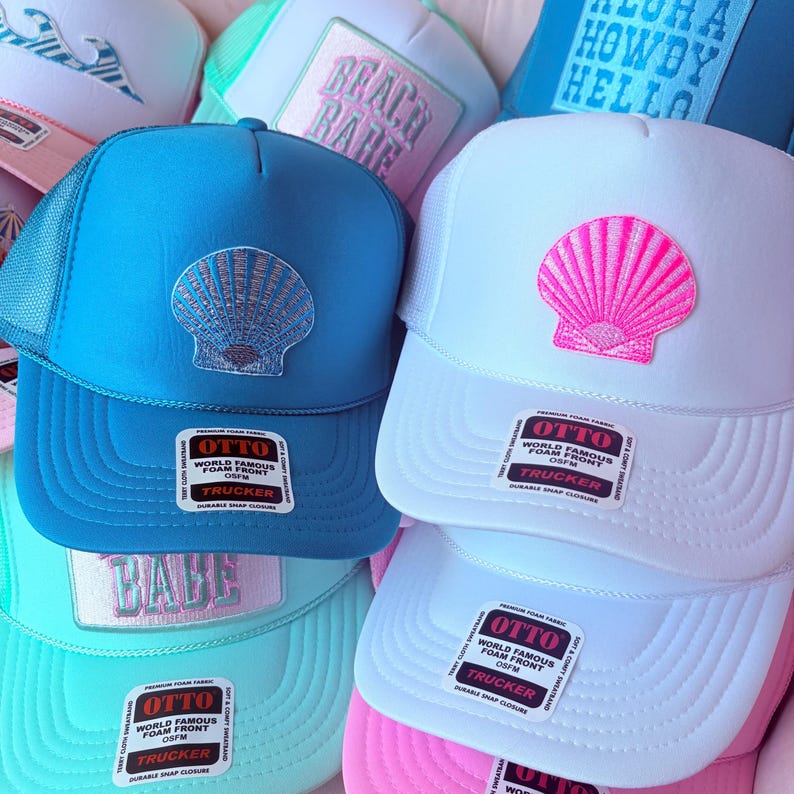 May include: A collection of trucker hats in various colors, including blue, white, and pink. Each hat features a seashell design on the front. The hats have a mesh back and a snap closure.