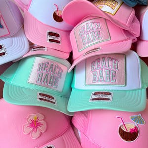 May include: A collection of trucker hats in various colors, including pink, white, and mint green. Each hat features a rectangular patch with the text "BEACH BABE" in a pastel color scheme. Some hats have additional embroidered details like a flower or a coconut drink.