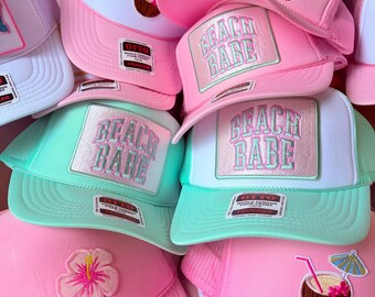 May include: A collection of trucker hats in various colors, including pink, white, and mint green. Each hat features a rectangular patch with the text "BEACH BABE" in a pastel color scheme. Some hats have additional embroidered details like a flower or a coconut drink.