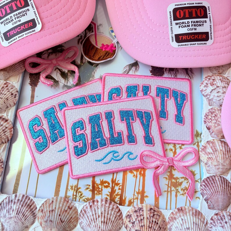 May include: Pink trucker hats with "OTTO" and "WORLD FAMOUS FOAM FRONT" labels. Patches with the word "SALTY" in blue glitter lettering are also visible. The image is decorated with seashells and pink bows, evoking a beach theme.