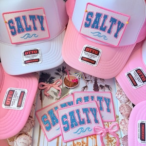 May include: White and pink trucker hats with the word "SALTY" in blue glitter lettering on a patch. The hats have a mesh back and a curved brim. The image also includes patches with the word "SALTY".