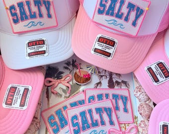 May include: White and pink trucker hats with the word "SALTY" in blue glitter lettering on a patch. The hats have a mesh back and a curved brim. The image also includes patches with the word "SALTY".
