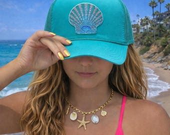 May include: A turquoise trucker hat with a seashell patch. The hat has a mesh back and a curved brim. A gold necklace with seashell charms and a starfish pendant is also visible.