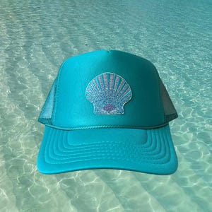 May include: A turquoise trucker hat with a seashell patch. The hat features a mesh back and a curved brim. The seashell design is light blue and white, set against a turquoise background. The hat is a summer accessory.