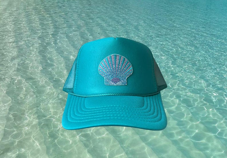 May include: A turquoise trucker hat with a seashell patch. The hat features a mesh back and a curved brim. The seashell design is light blue and white, set against a turquoise background. The hat is a summer accessory.