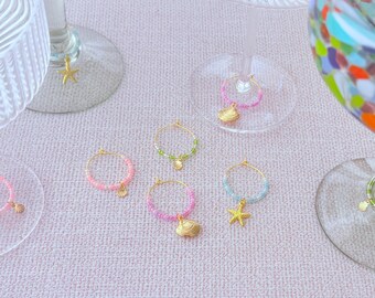 May include: Gold-toned wine glass charms with colorful beads and seashell, starfish, and shell pendants. The charms are arranged on a light pink surface, near wine glasses.