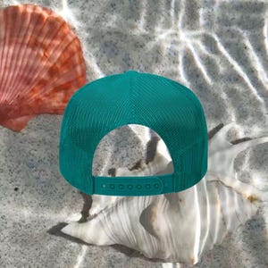 May include: A teal trucker hat with a mesh back sits on a sandy surface, next to a large white seashell and a reddish-orange scallop shell. The hat has an adjustable snap closure.