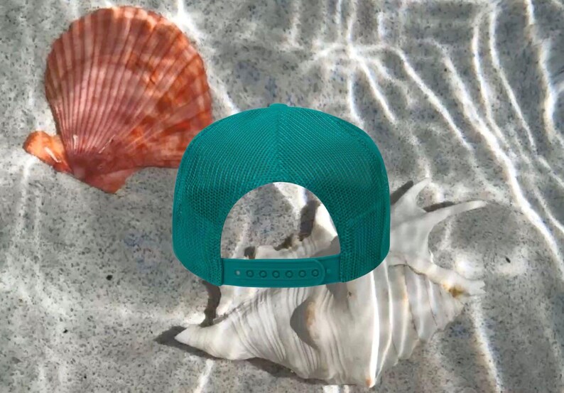 May include: A teal trucker hat with a mesh back sits on a sandy surface, next to a large white seashell and a reddish-orange scallop shell. The hat has an adjustable snap closure.