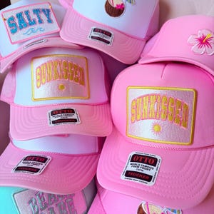 May include: A collection of pink trucker hats with white mesh backs. Some hats feature the text "SUNKISSED" in gold lettering, while others say "SALTY" in blue. Each hat has a "OTTO" brand tag.