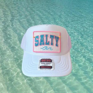 May include: White trucker hat with a foam front. The hat features a rectangular patch with the word "SALTY" in turquoise letters outlined in pink. Below the word "SALTY" is a small turquoise wave. The hat has a mesh back and a snap closure.