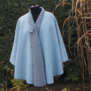 Op de afbeelding: Een lichtblauwe cape met een grijze voering. De cape is gemaakt van een zacht, pluche materiaal en heeft een eenvoudig, elegant ontwerp.